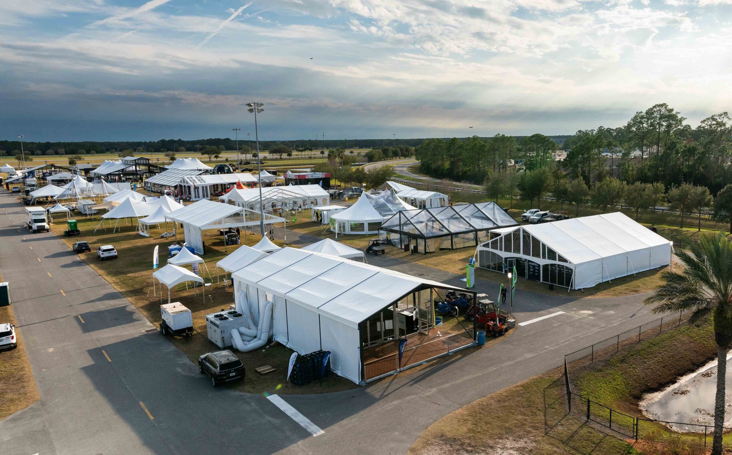 Aerial view of a tent exhibition area featuring multiple white tents, vehicles, and a landscape of greenery under a cloudy sky.