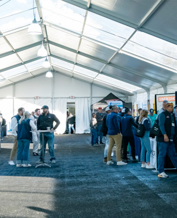 A spacious tent filled with people engaging in discussions, with booths in the background and bright overhead lights illuminating the scene.