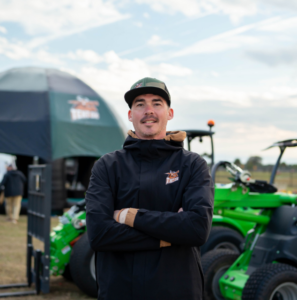 Andy Mattrick Man with a relaxed smile stands with arms crossed, wearing a black jacket and cap, against a tent and green machinery in an outdoor setting.