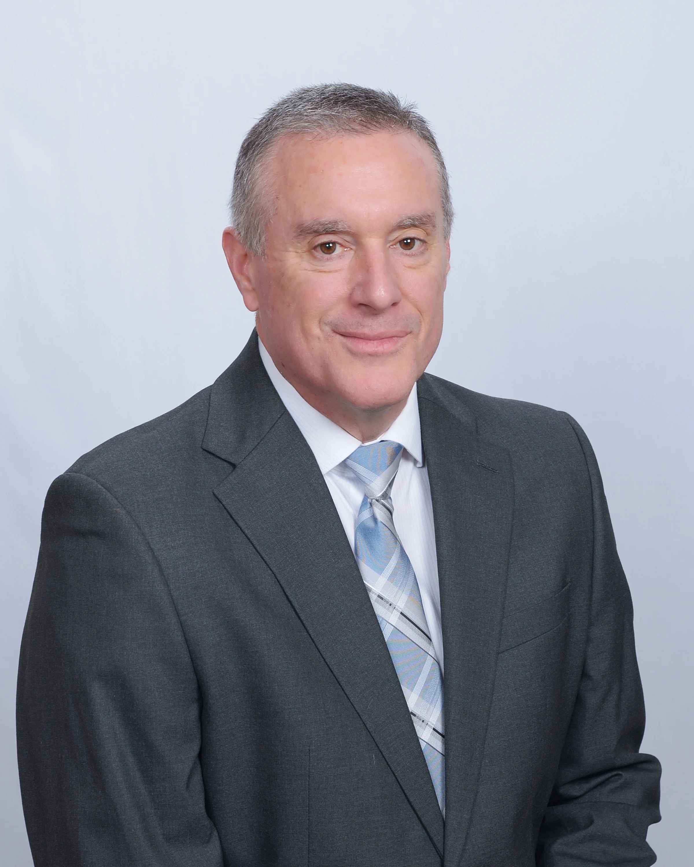 Professional headshot of a man in a dark grey suit with a light blue and white plaid tie, set against a plain white background.