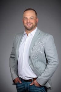 Nate Niespodziany Portrait Photography by Dan Henry / DanHenryPhotography.com Smiling man wearing a light gray blazer and white shirt, with hands in pockets, against a muted gray background.