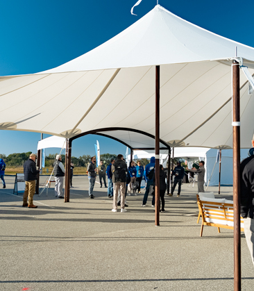 A spacious white pole tent with guests mingling underneath, set against a clear blue sky and a concrete surface.