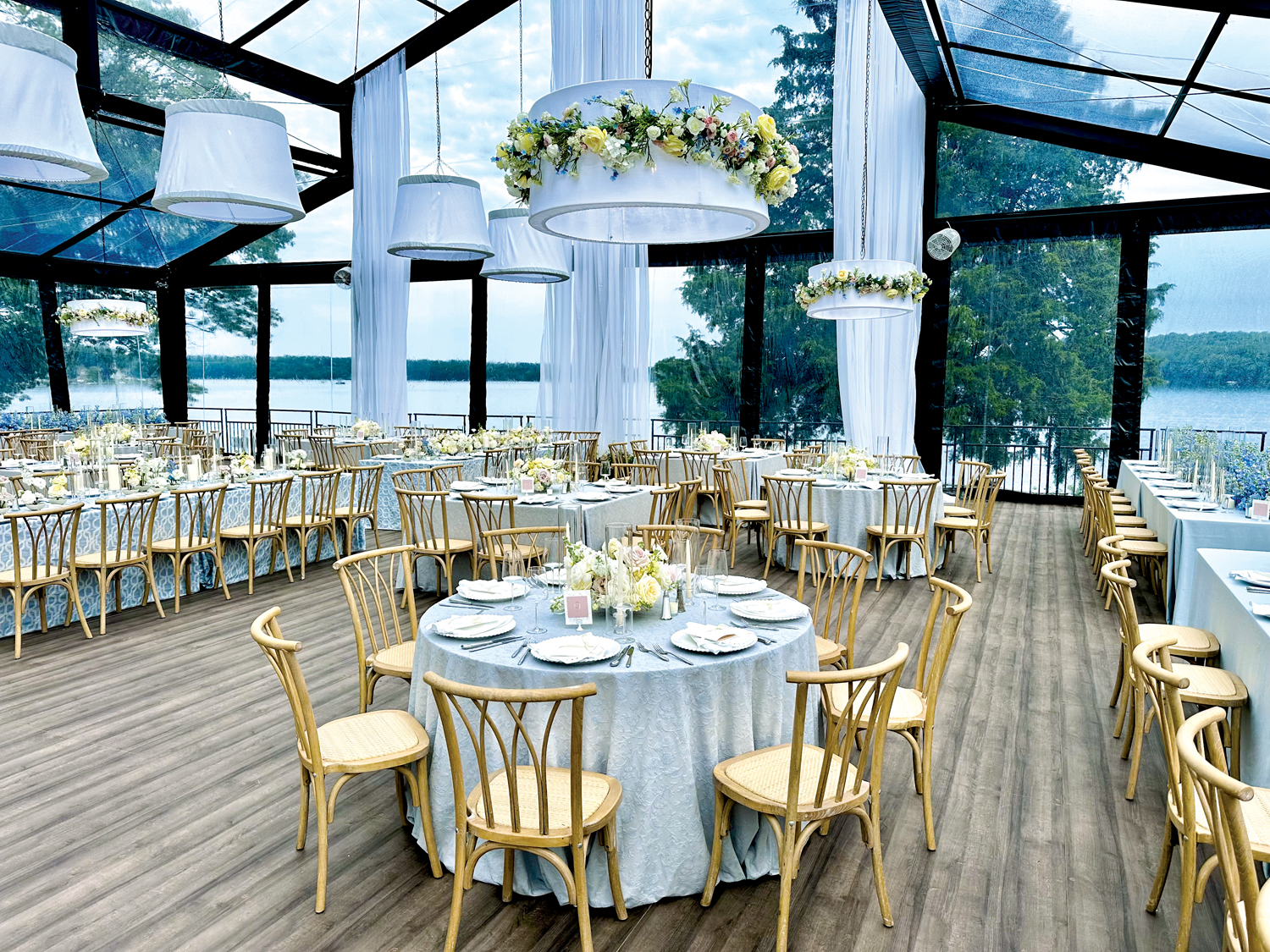 Elegant dining setup in a glass pavilion overlooking a lake, featuring round tables, floral centerpieces, and wood accents.