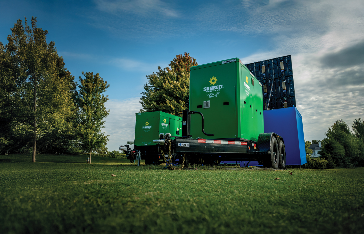 Green utility trailers are parked on grass, surrounded by trees and a blue structure, under a partly cloudy sky.