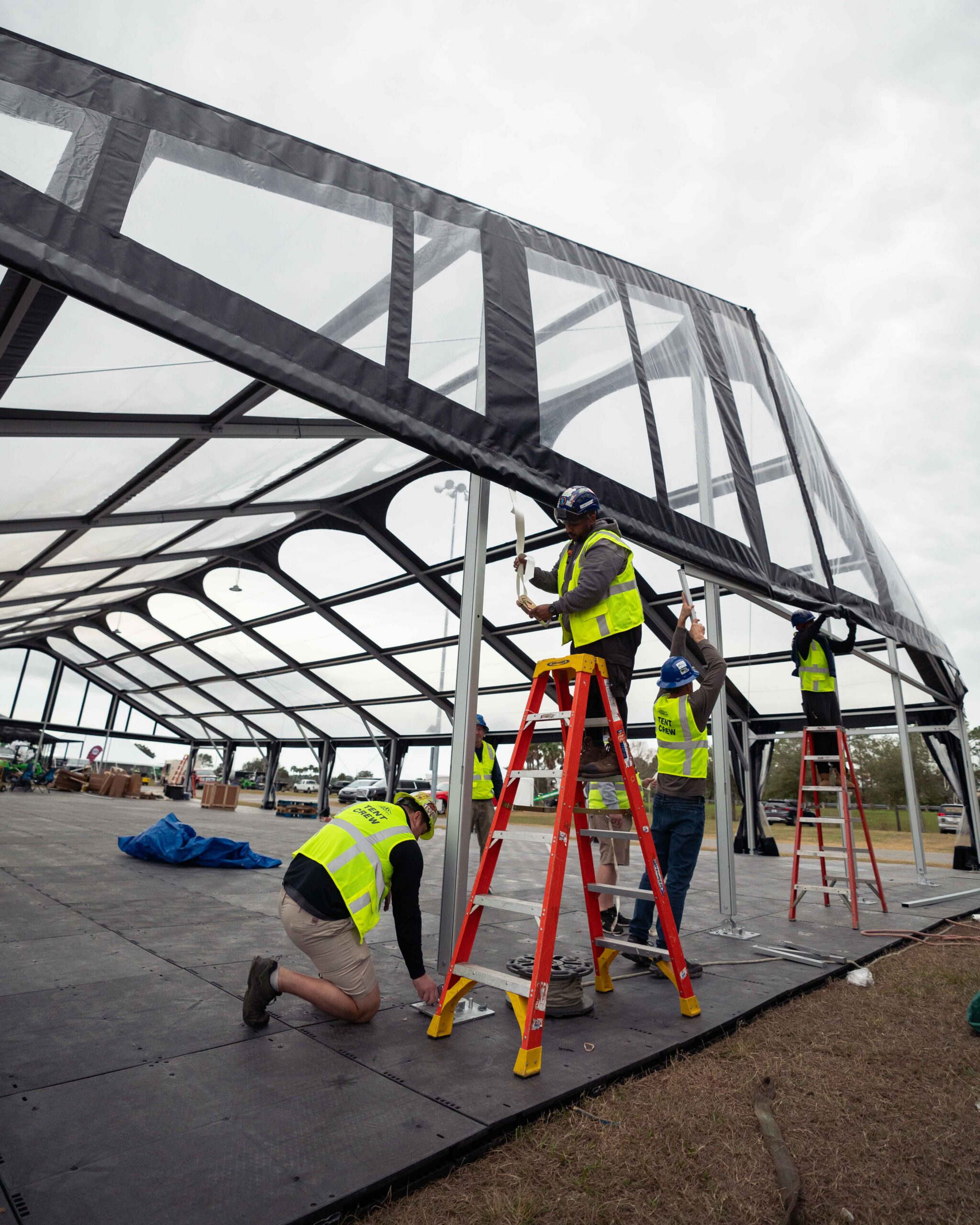 Anchor-2 Workers in neon vests set up a large tent using ladders, with a cloudy sky overhead and equipment scattered on the ground.