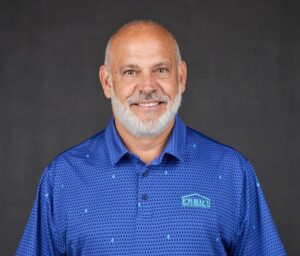 BrianStumph_HeadShot Smiling man with a beard wearing a blue polo shirt with a logo, set against a dark gray background.