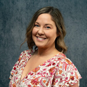 Camile Fox – Headshot Smiling woman with shoulder-length brown hair, wearing a floral-patterned blouse in front of a textured gray background.