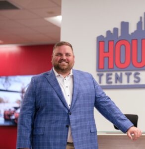 HTE A man in a blue checked blazer smiles while standing next to a reception area with a red and white sign that reads "HOUSE TENTS."