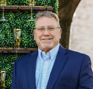 Headshot_Mike Holland Man with glasses and a neatly groomed beard smiles, dressed in a navy blazer over a light checkered shirt, against a greenery backdrop.