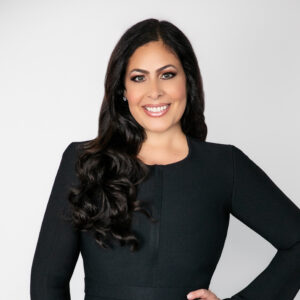 Stacie House_Web_2 Smiling woman with long, wavy dark hair wearing a fitted black dress, posed against a light gray background.