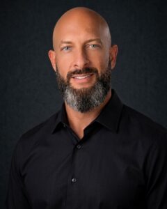 steven herring headshot A man with a bald head and a full beard, wearing a black shirt, smiles against a dark textured background.